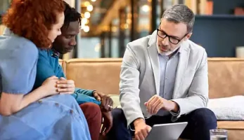 image: Man in business casual clothing speaking to a couple, pointing to a tablet