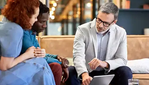 image: Man in business casual clothing speaking to a couple, pointing to a tablet