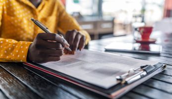 image: Person holding a pen over a clipboard filled with documents that must be filled out