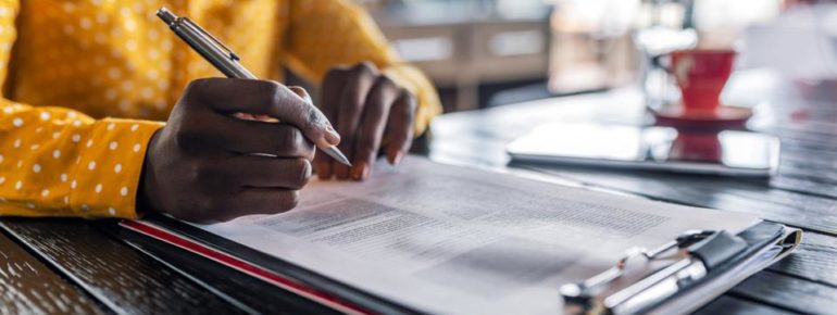 image: Person holding a pen over a clipboard filled with documents that must be filled out