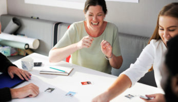 image: Group of individuals, sitting at a table, laughing as they look at polaroid pictures