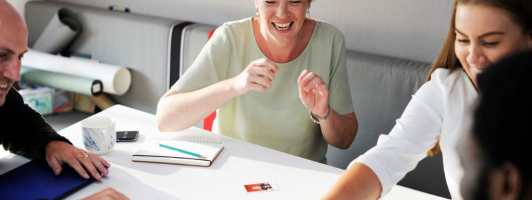 image: Group of individuals, sitting at a table, laughing as they look at polaroid pictures