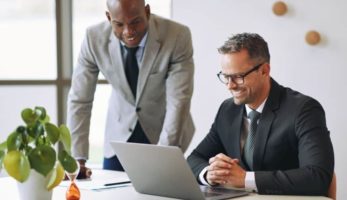 image: Businessman sitting at a desk with a suit and tie, smiling at someone via a video call, as a colleague (also in a suit) watches over