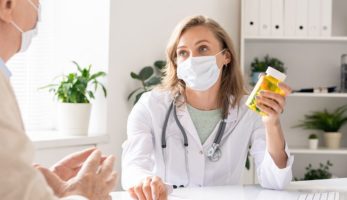 image: Doctor wearing a mask, holding a bottle of pills, speaking to a patient