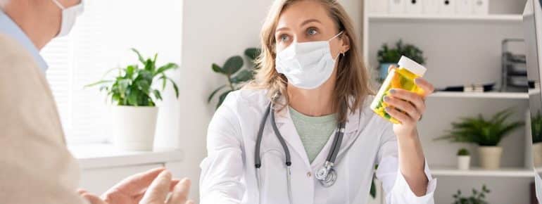 image: Doctor wearing a mask, holding a bottle of pills, speaking to a patient